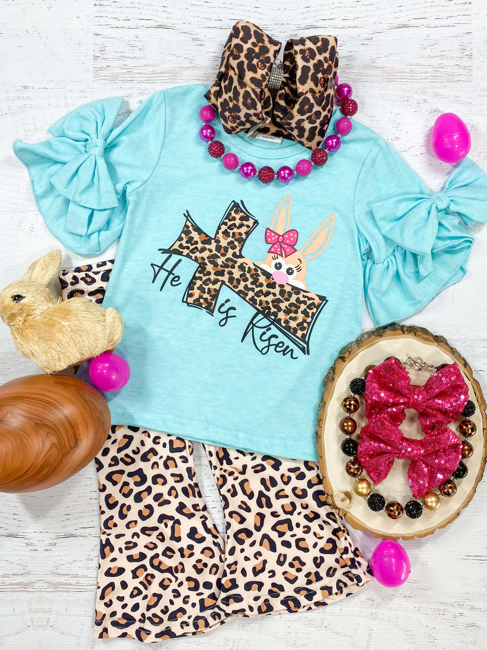 He Is Risen Black lettering with animal print cross and peek-a-boo bunny, blue top with ruffle sleeves and bows paired with animal print shorts.